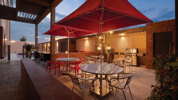 Een terras buiten met ronde tafels en stoelen, een rode parasol en een barbecue.