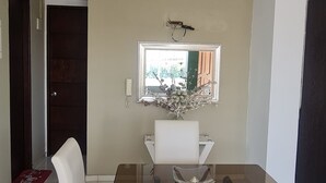 A dining area with a glass-top table, white cushioned chairs, a vase with flowers, and a framed mirror on the wall. - Lindo y Acogedor Apartamento Cerca de Todo (Barranquilla)