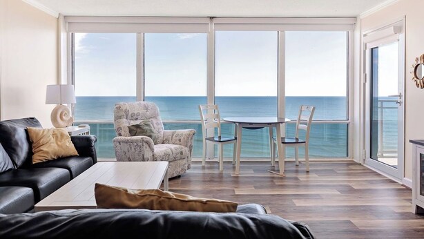 Una sala de estar con un ventanal grande, un sofá de cuero negro, un sillón estampado, una mesita y vista al mar.