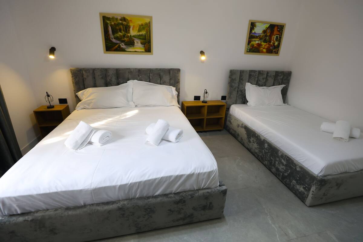 A hotel room with two beds, each with white linens and towels. There are two nightstands with lamps and a small shelf. The walls are plain with two framed pictures.