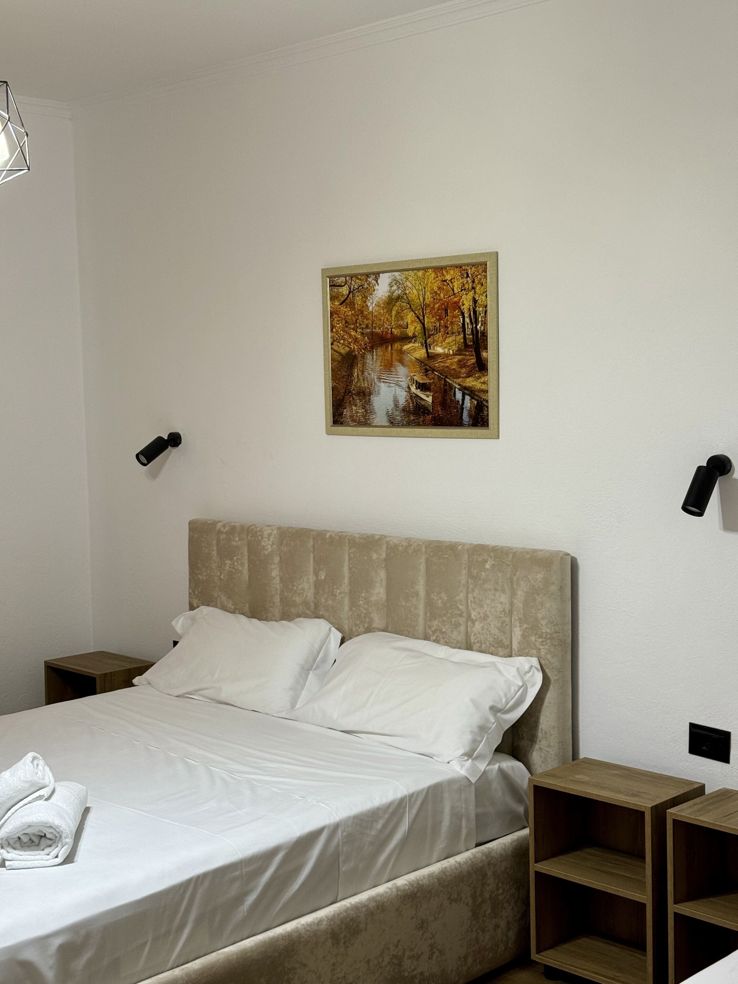 A bedroom with a bed, two bedside tables, a nightstand, a painting of a river scene, and a wall-mounted telephone.