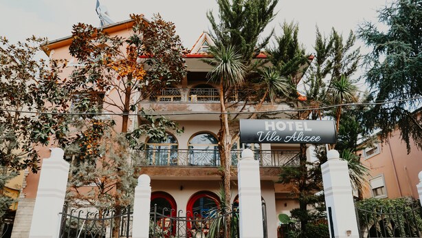 Un hotel con un letrero que dice "HOTEL Vila Huzee". Cuenta con una estructura de dos pisos con balcones, puertas rojas y una variedad de vegetación, incluyendo árboles y plantas en macetas.