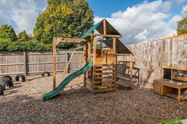 A wooden play structure with a slide and climbing elements, surrounded by a wooden fence and trees. - Hot tub, games room & cinema in Bournemouth (Bournemouth)