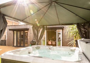 A hot tub under a covered patio with a wooden deck, a glass door leading to a patio area, and a table with chairs outside. - Hot tub, games room & cinema in Bournemouth (Bournemouth)