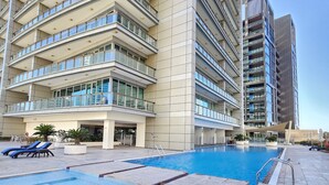A modern hotel with a large outdoor swimming pool, multiple balconies, and a clear sky. - Vacay Lettings - 8 Boulevard Walk (Dubai)