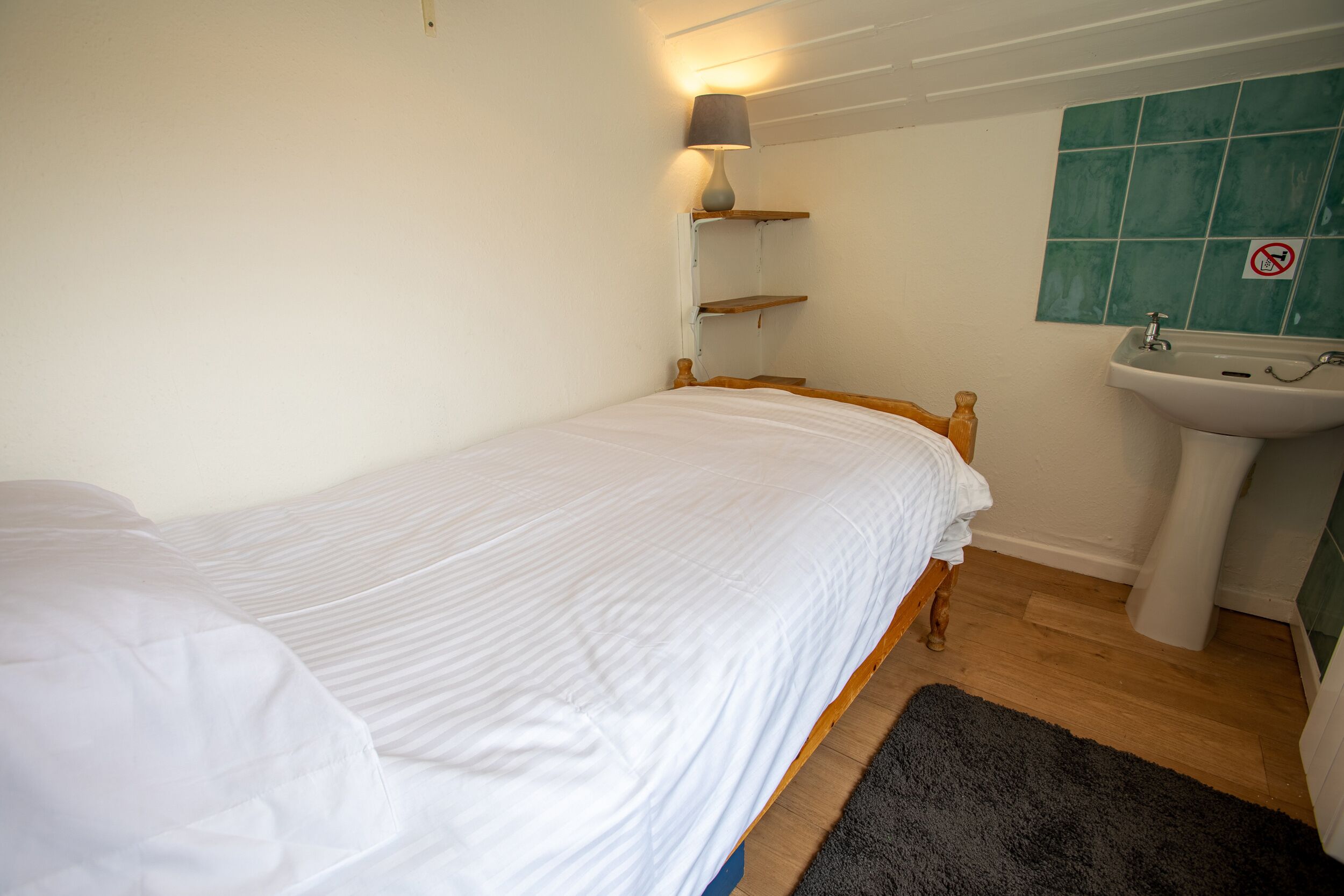 Een kleine, schone kamer met een enkel bed, een nachtkastje, een lamp, een plank en een wastafel.