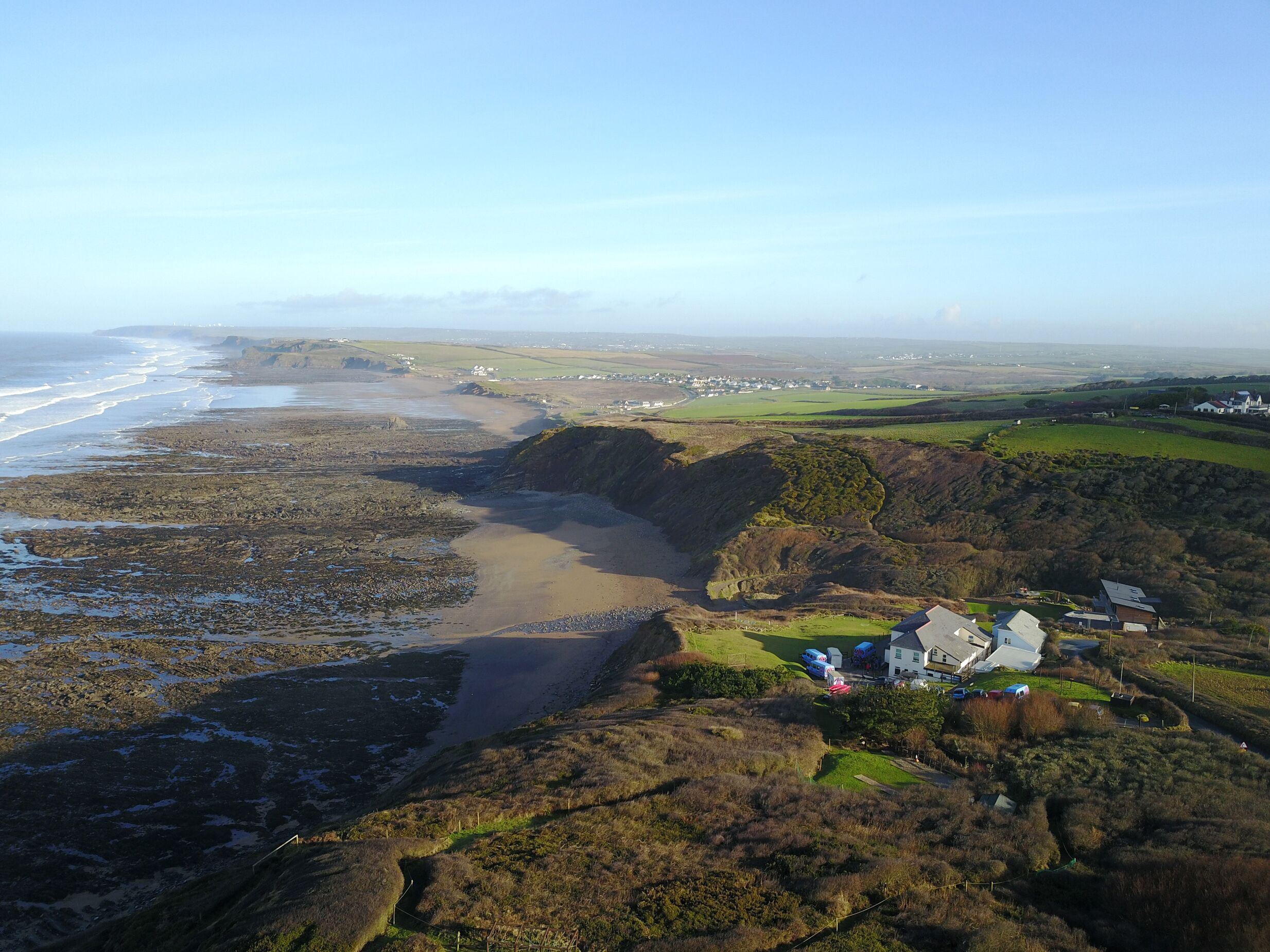 An aerial view of a coastal area with a rocky shoreline, a small settlement, and green fields.