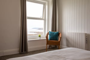 A hotel room with a bed, a chair with a pillow, a window with curtains, and a small plant on the windowsill.