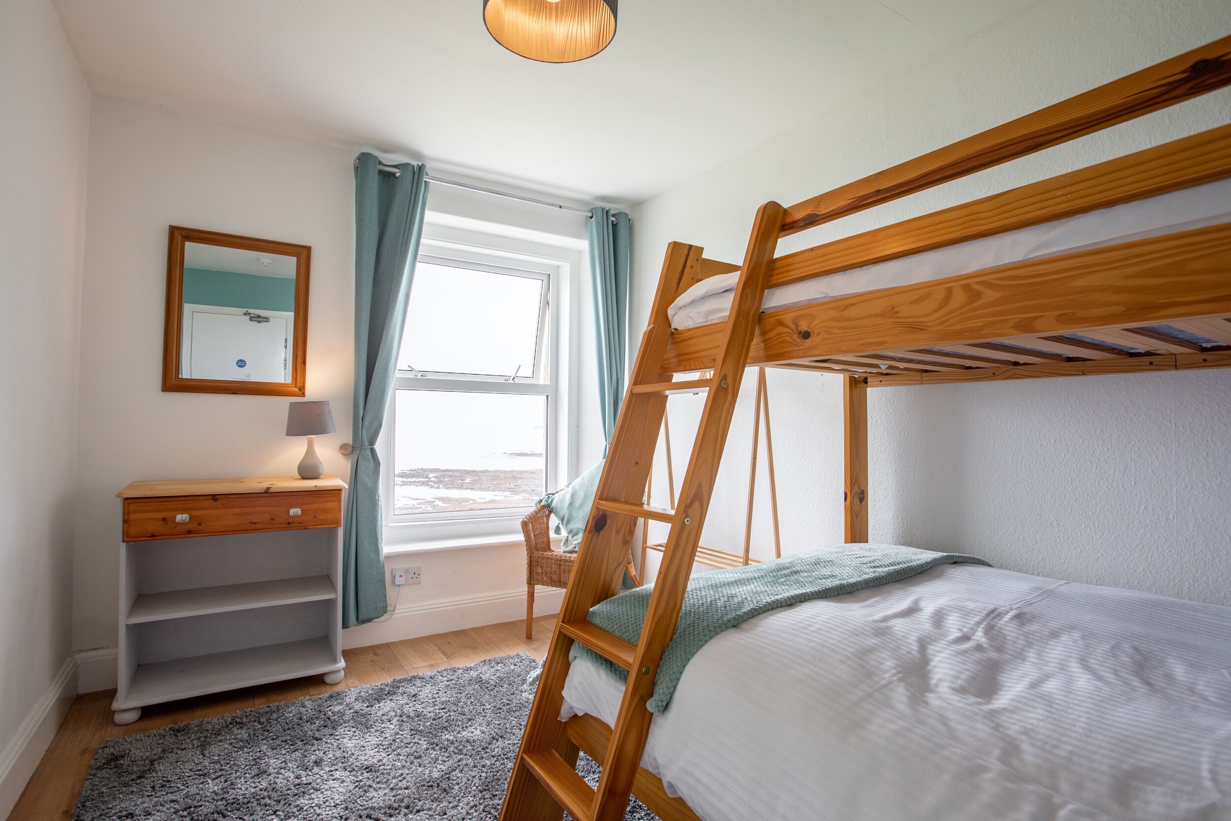 A bunk bed room with a wooden ladder, a desk with drawers, a chair, and a window with curtains.