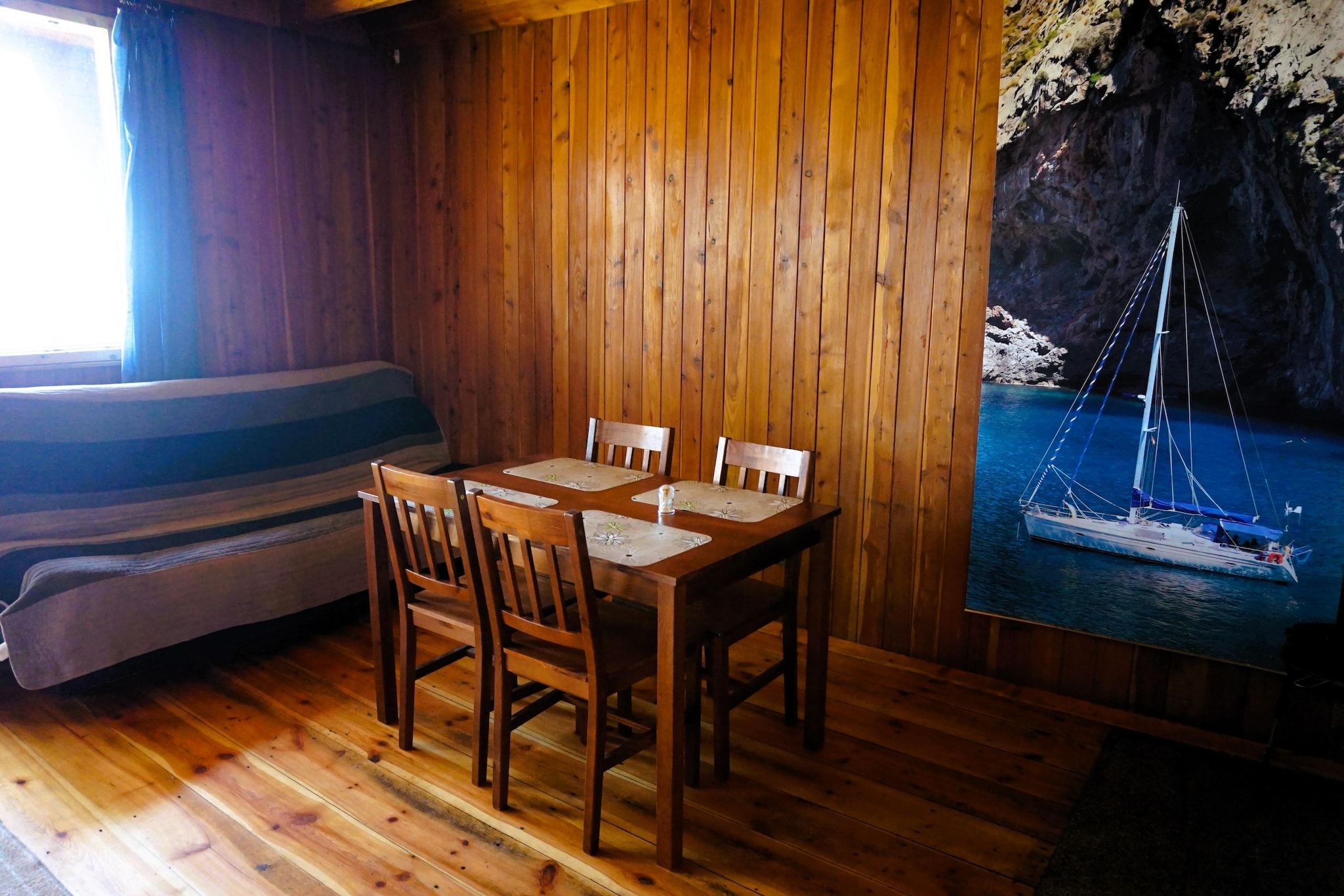 A wooden interior with a dining table and chairs, a sofa, and a mural of a sailboat on a cliff.