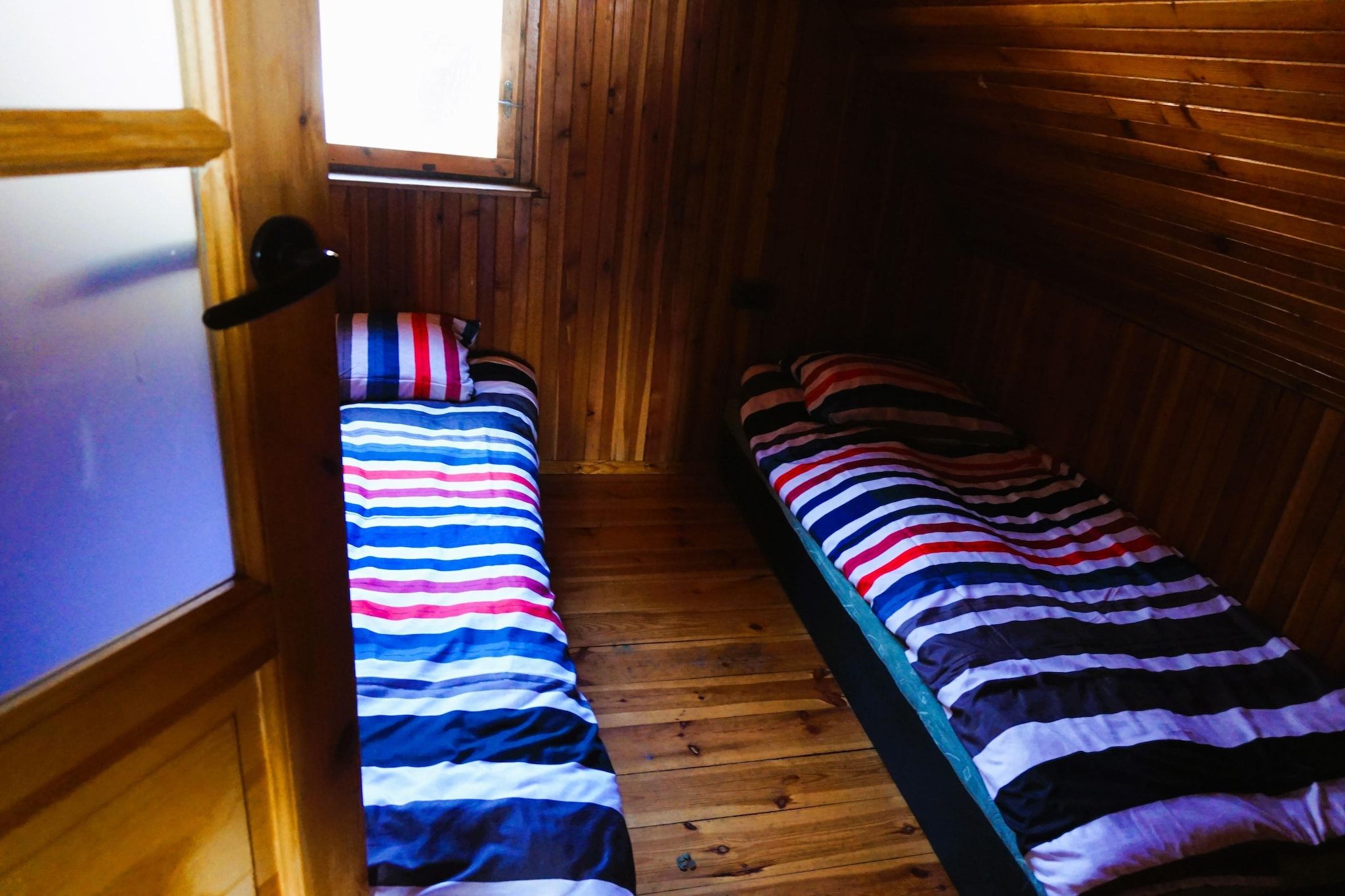 A narrow room with two beds aligned against the wall, wooden flooring, and a window at the far end.