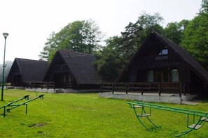 A row of rustic, triangular-roofed cabins with a grassy area in front, benches, and a lamppost. - Summer House in Łazy by Lake Jamno (Łazy)