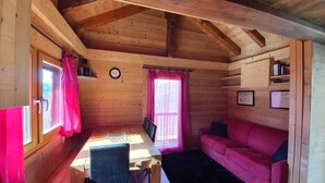 A wooden cabin interior with a dining table, chairs, a pink sofa, and a window with curtains.