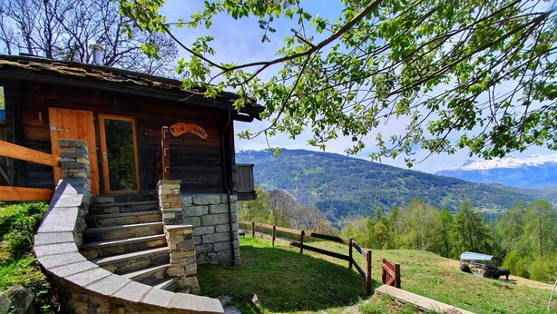 A rustic cabin with a stone staircase leading to a wooden door, surrounded by a fenced area with a view of mountains.