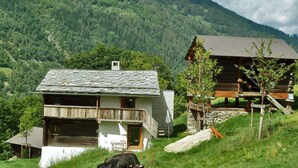 A rustic mountain cabin with a stone roof, surrounded by lush greenery and a cow grazing nearby.
