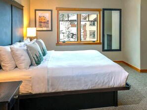 A neatly made bed with white linens, a bedside table with a lamp, a window with a view of buildings, and a mirror on the wall.