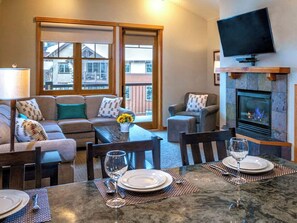 A living room with a sofa, chairs, a dining table set for two, a fireplace, and a TV mounted above it.
