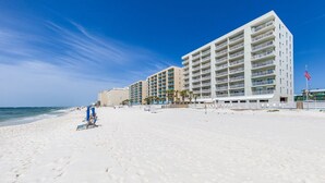 Un hotel frente a la playa con varios pisos, una playa de arena en primer plano y un cielo azul despejado.