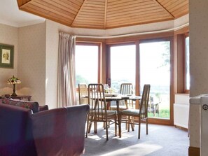Una habitación con una mesa de comedor y sillas, un sofá, una lámpara y vistas a zonas verdes a través de ventanas amplias.