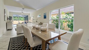 Un comedor moderno con una mesa de madera y sillas blancas, ventanas amplias con vistas a zonas verdes y un ventilador de techo.