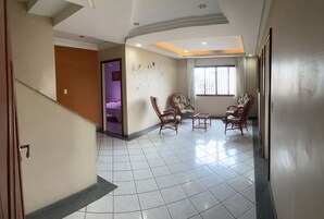 Uma sala ampla com piso ladrilhado, uma escada e um espaço de estar com cadeiras de vime e uma mesinha.