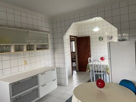 Cozinha com paredes revestidas de azulejos brancos, porta de madeira, frigorífico branco e mesa de jantar com cadeiras.