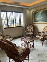 Quarto com mobiliário de vime, uma pintura e vista para edifícios através de grandes janelas.