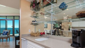 A kitchen with a glass shelf holding decorative items, a coffee maker, and a view of the ocean through a sliding door.