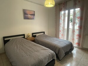 Une chambre avec deux lits, une table de chevet avec une lampe, un tableau au mur et une fenêtre avec des rideaux à motifs floraux.