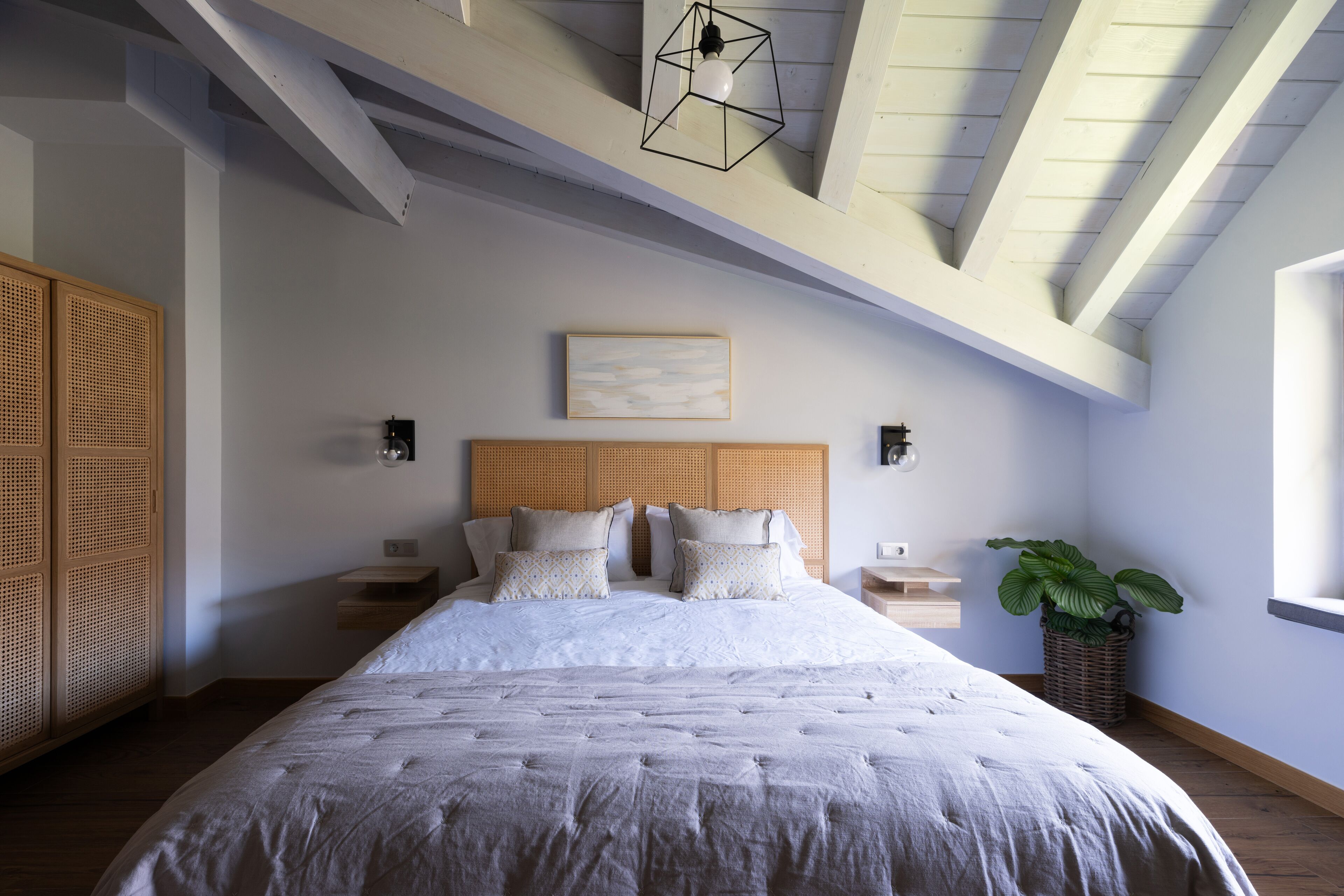 A bedroom with a large bed, wooden headboard, bedside tables, a wall-mounted light fixture, and a potted plant.