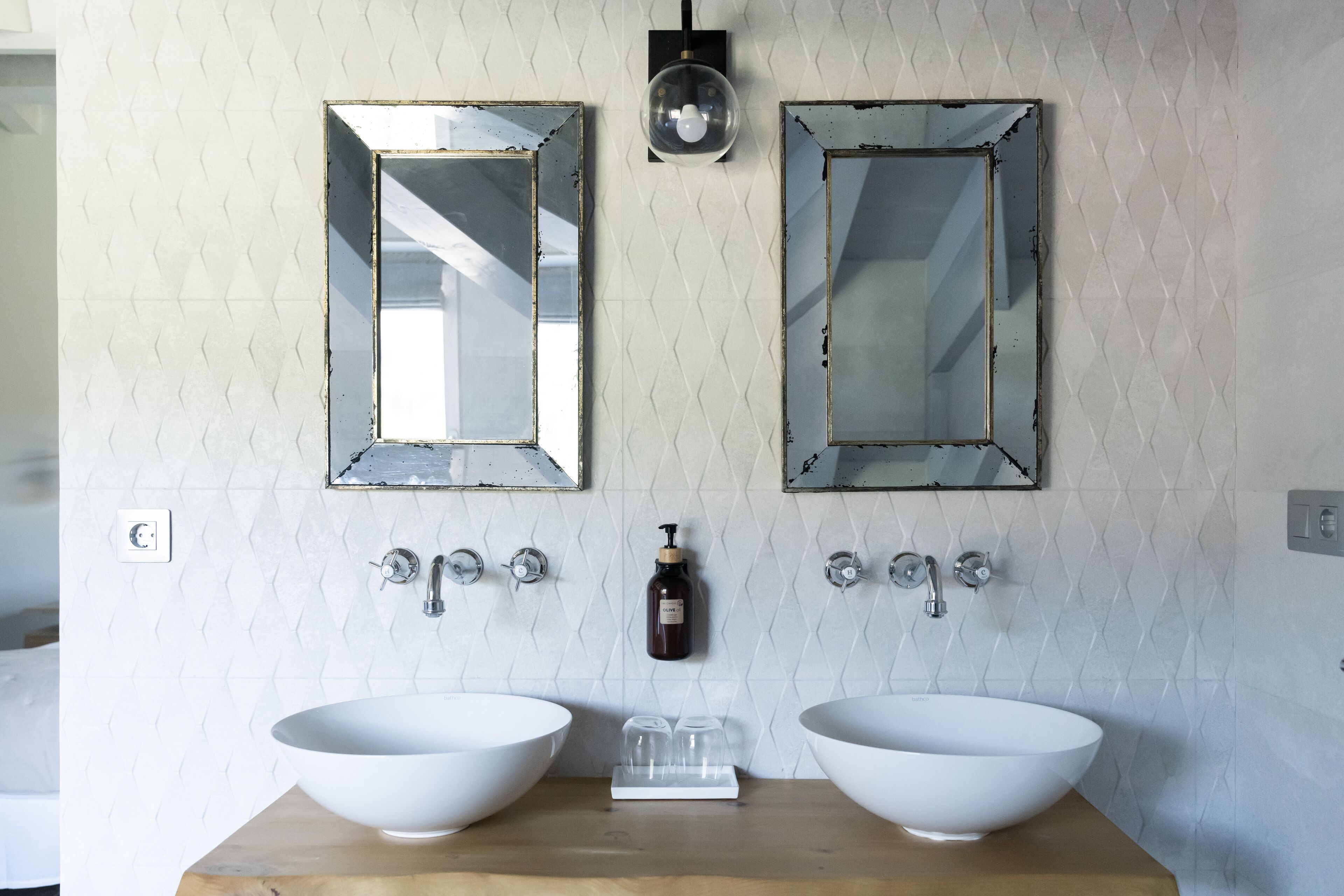 Un baño con dos lavamanos blancos, una encimera de madera, dos espejos, un aplique de pared y un dispensador de jabón.