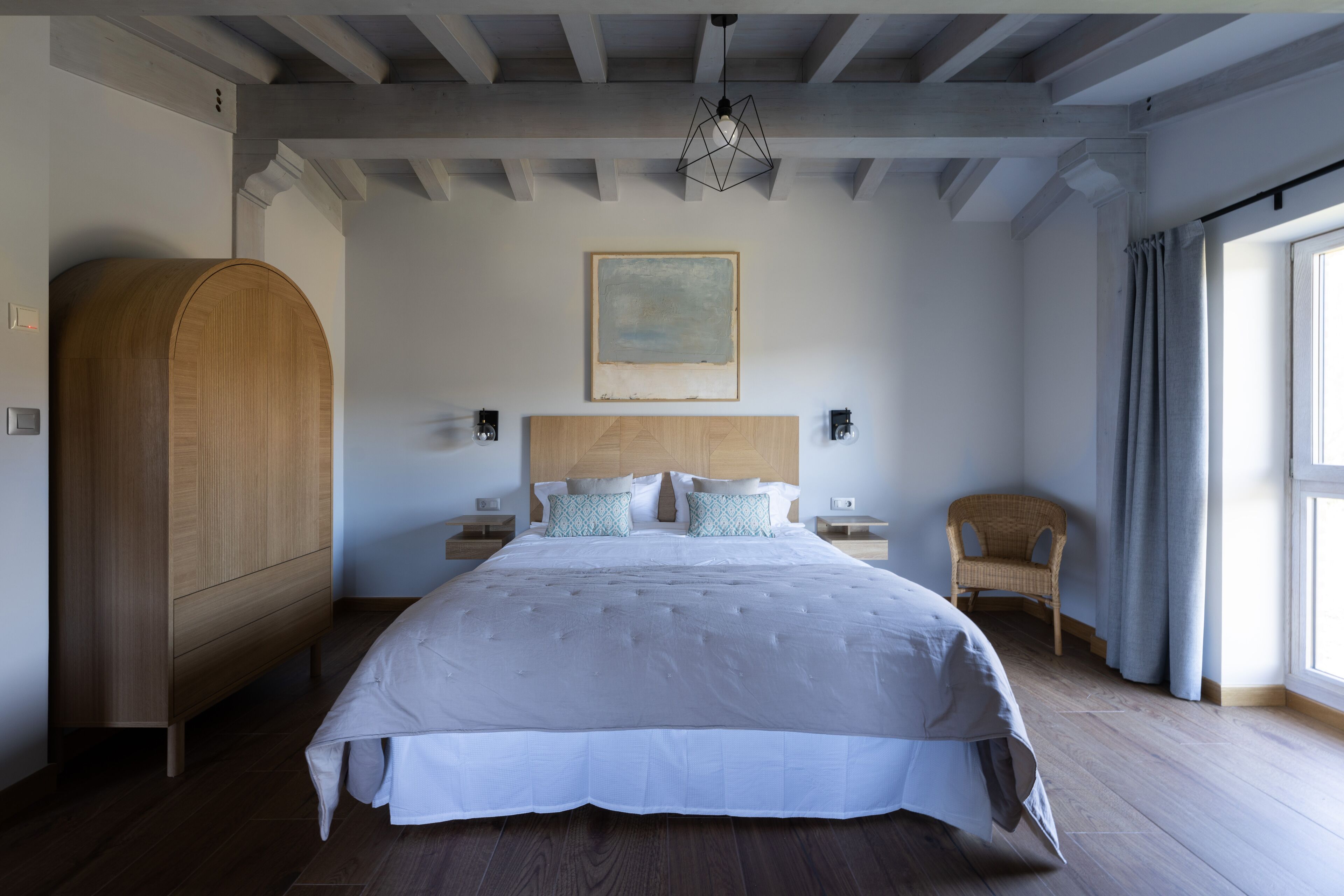 Een slaapkamer met een groot bed, een houten garderobe, twee nachtkastjes, een aan de muur bevestigd schilderij en een raam met gordijnen.