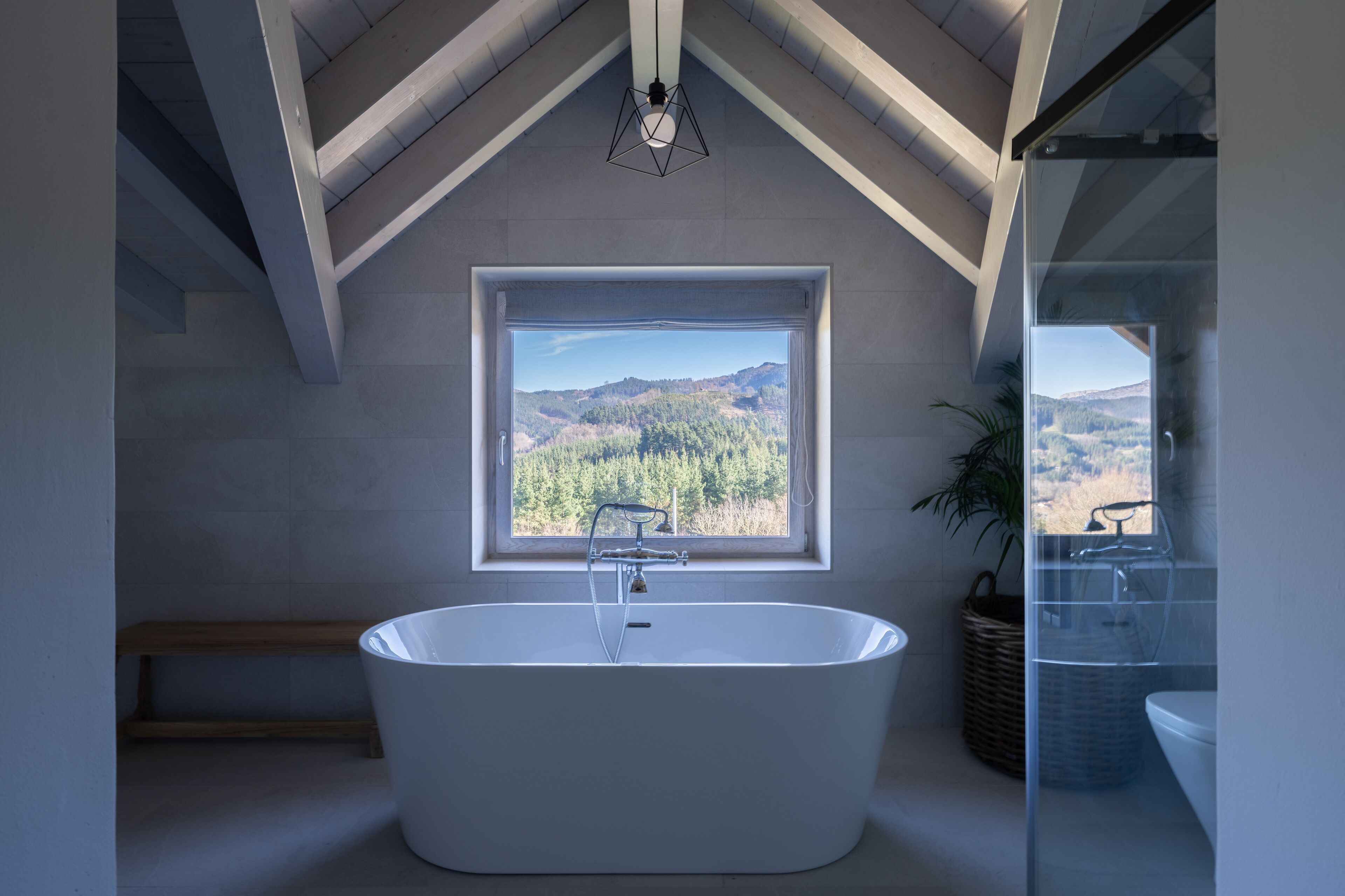Une salle de bain moderne avec une baignoire sur pieds, une grande fenêtre donnant sur un paysage de montagnes boisées et une lucarne au plafond.