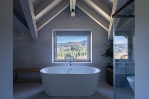 A modern bathroom with a freestanding bathtub, a large window offering a view of a forested mountain range, and a skylight above. - Soloandie (Zeanuri)