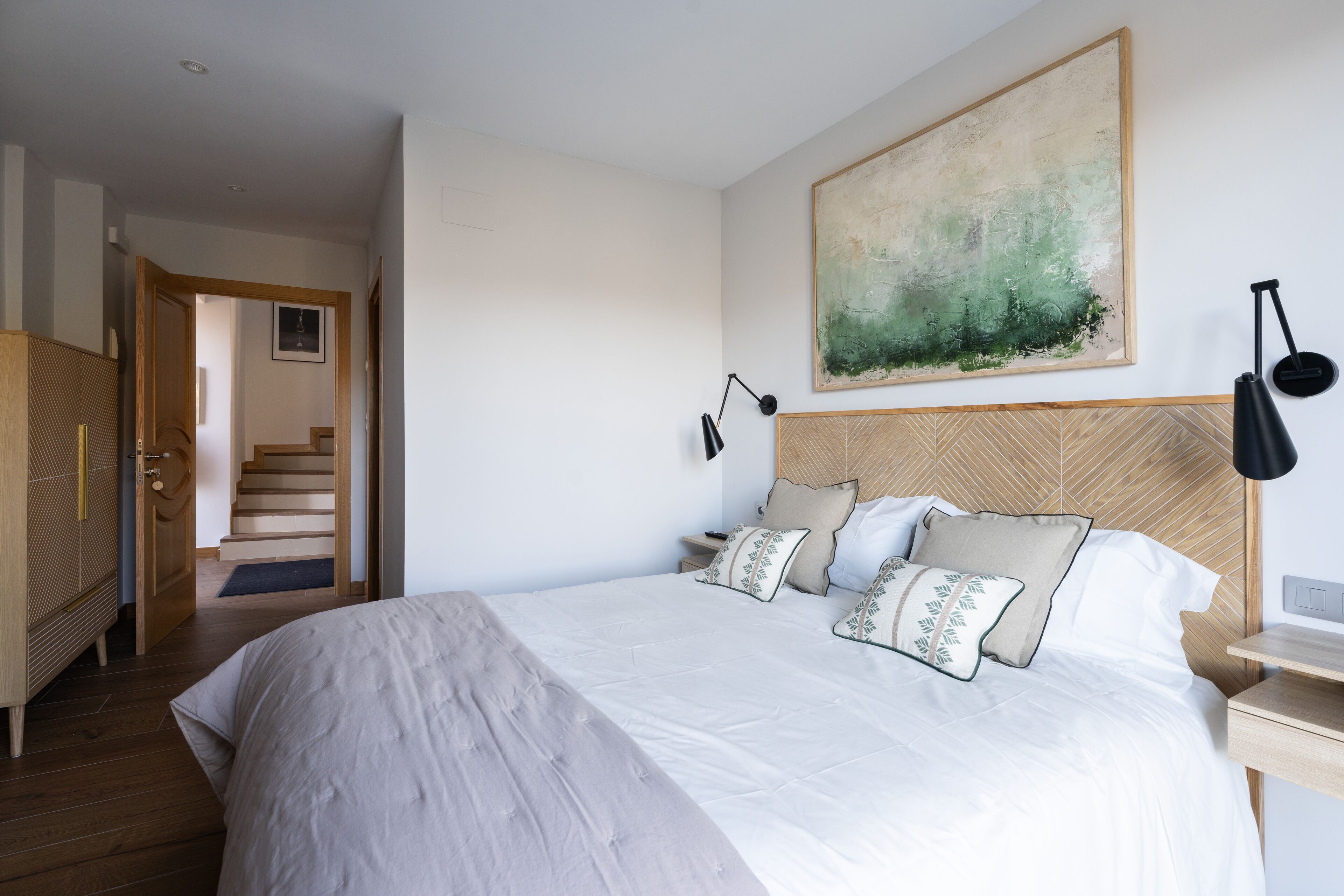 A bedroom with a large bed, a wooden headboard, a nightstand, a wall-mounted lamp, and a painting.