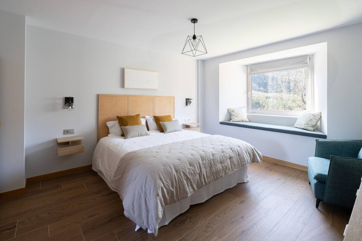 A bedroom with a large bed, a window with a view, a bedside table, and a chair.