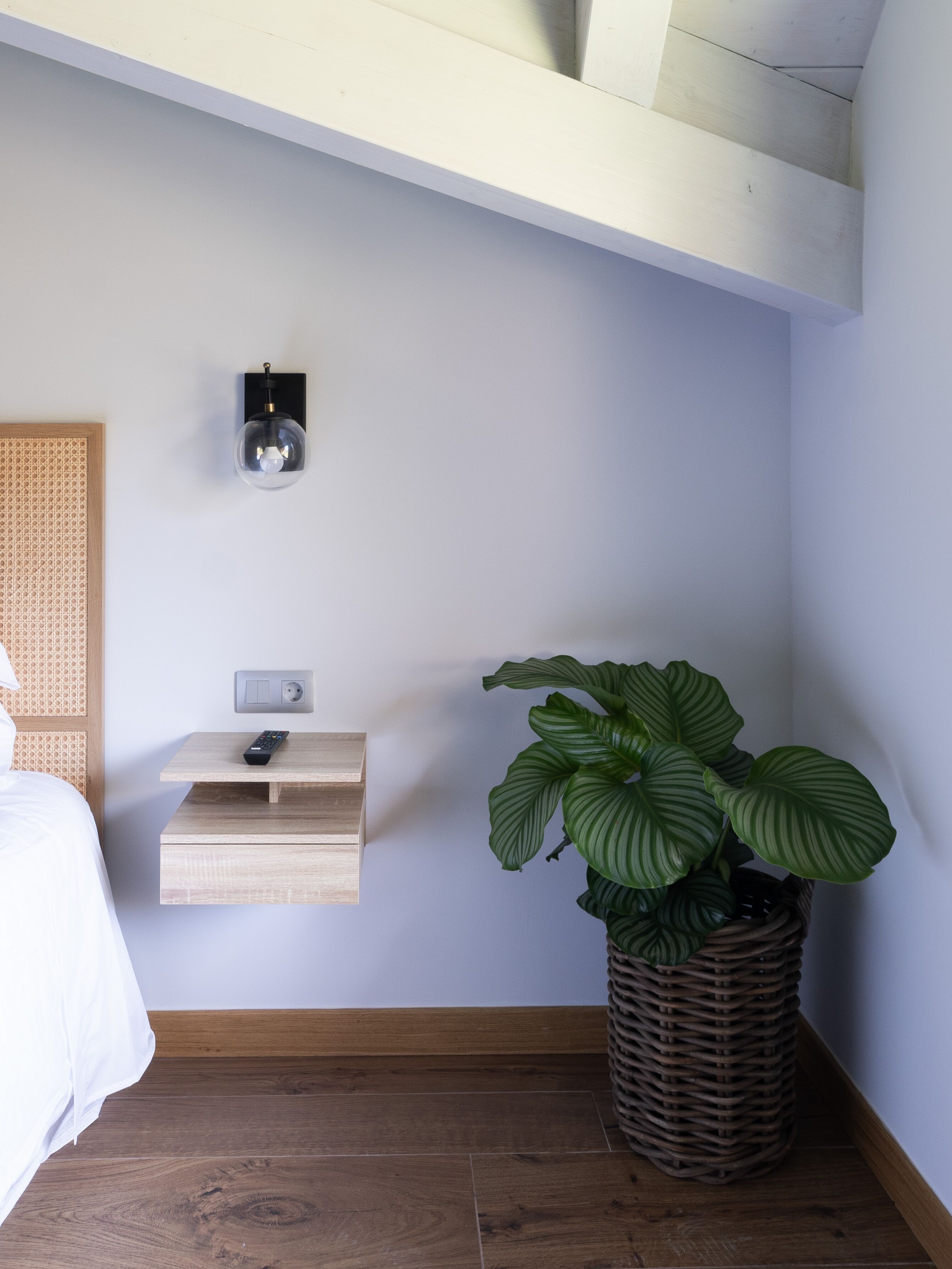 Een slaapkamer met een bed, een nachtkastje met een afstandsbediening, een wandlamp en een potplant.
