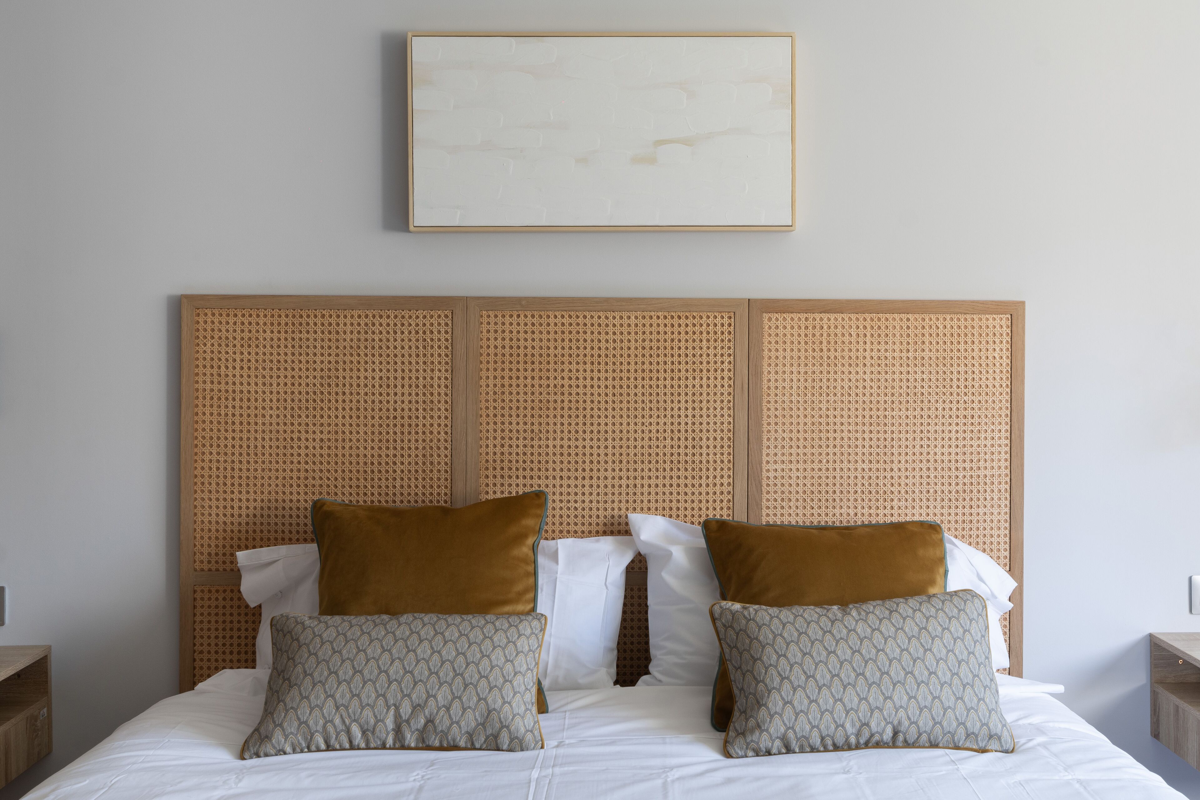 Un lit bien fait, avec deux oreillers, une tête de lit ornée d’un motif tissé et un tableau encadré au-dessus.
