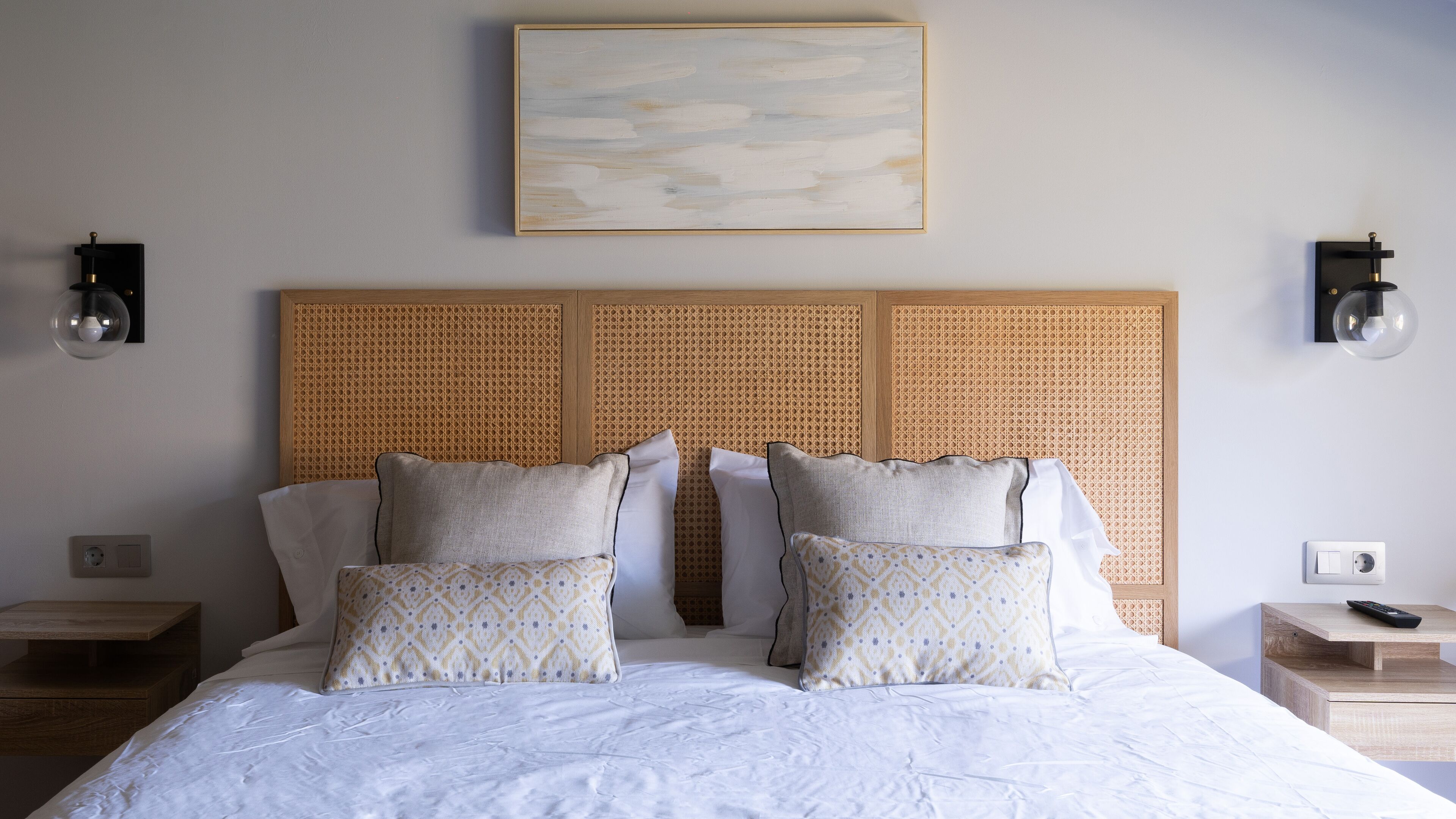 Una cama bien hecha con edredón blanco, dos almohadas con estampado geométrico y un cabecero tejido con textura. Hay dos mesitas de noche, cada una con una lámpara y un control remoto.