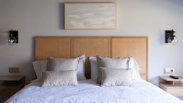 Una cama bien hecha con edredón blanco, dos almohadas con estampado geométrico y un cabecero tejido con textura. Hay dos mesitas de noche, cada una con una lámpara y un control remoto.