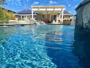 Une piscine à l’eau claire, une maison dotée d’un patio couvert et une cascade décorative.