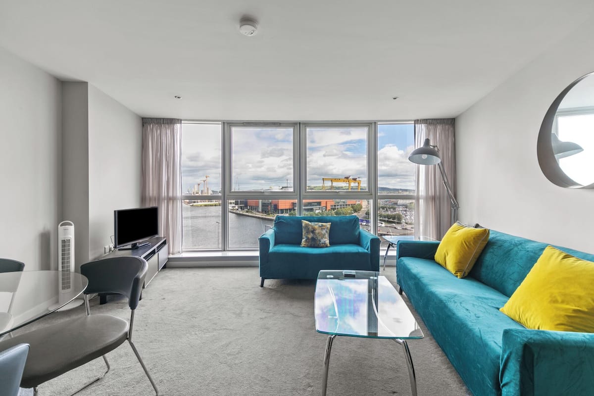 Un salon moderne avec une grande fenêtre, un canapé de couleur turquoise, une table basse en verre et un téléviseur.