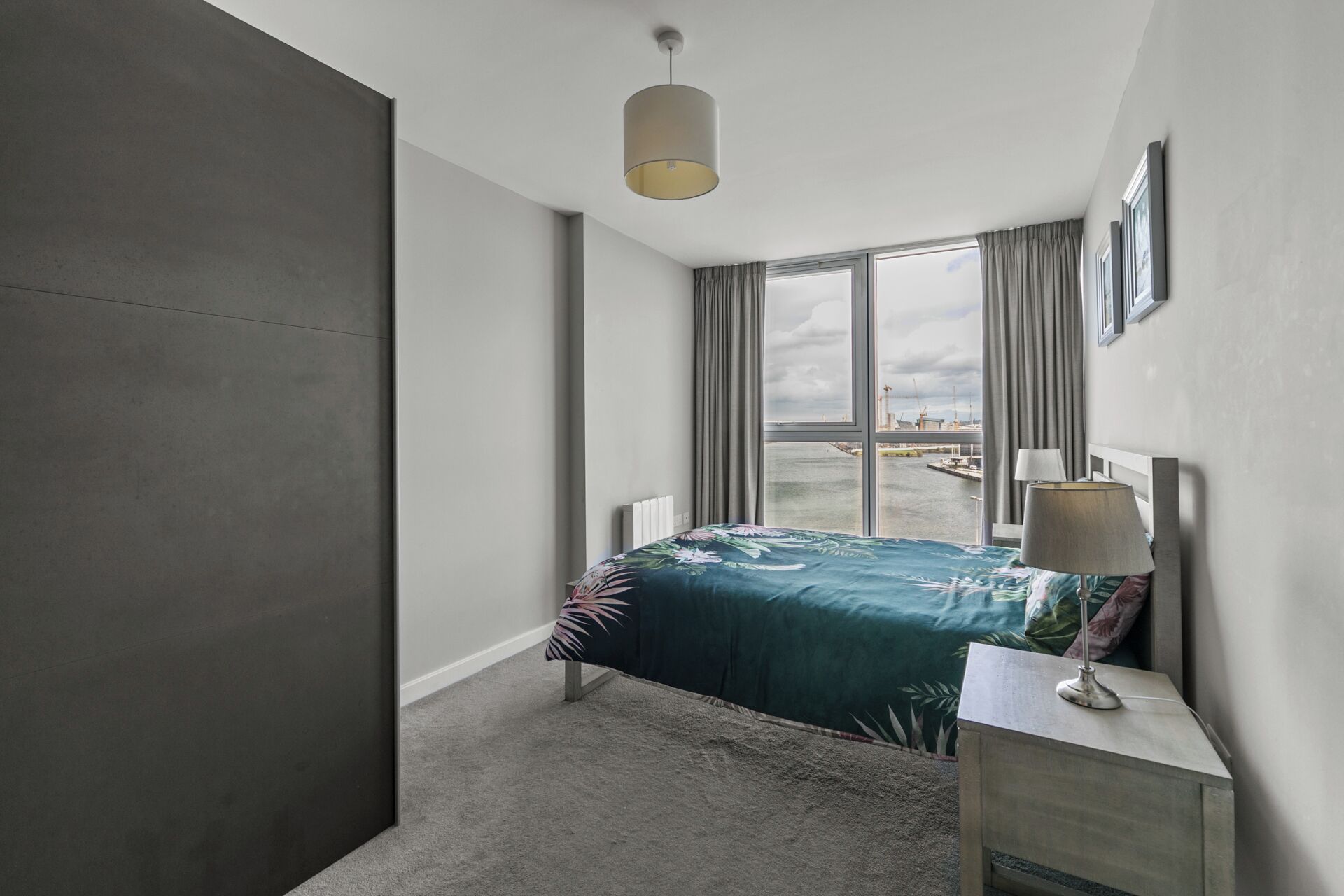 A modern hotel room with a large window, a bed with a floral bedspread, a bedside table with a lamp, and a view of the cityscape.