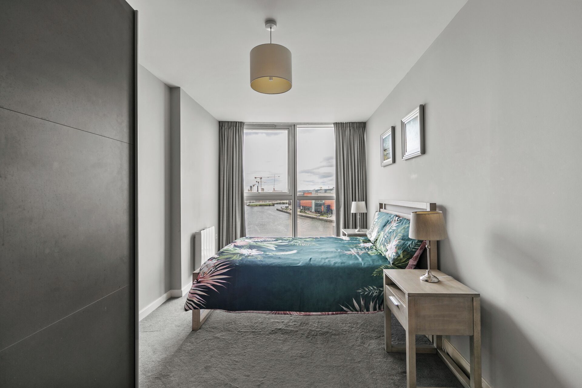 Une chambre à coucher moderne avec une grande fenêtre, un lit recouvert d’une couvre-lit à motifs floraux, une table de chevet avec une lampe, et une vue sur le paysage urbain.