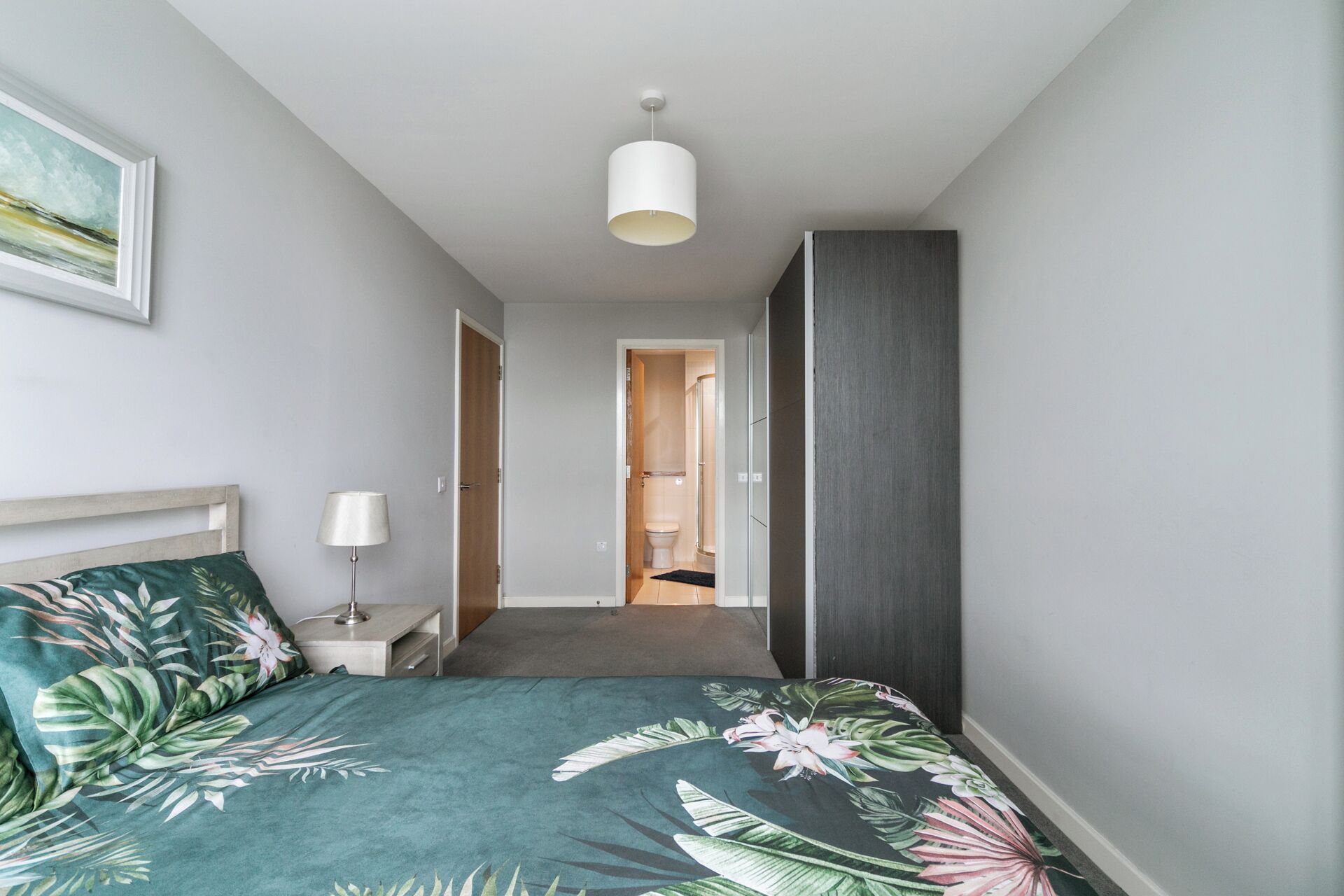 Habitación de hotel con cama, mesita de noche, lámpara, un cuadro y baño a la vista a través de una puerta abierta.