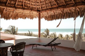 A beachside patio with a thatched roof, a lounge chair, and a view of the ocean.