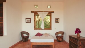 Quarto com cama, duas cadeiras de vime, cômoda de madeira e janela com vista para árvores.