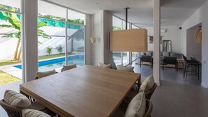 Una habitació espaiosa amb un gran taulell de fusta i cadires de vimet, grans finestres que donen a una piscina i vegetació.