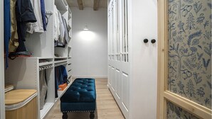 A narrow hallway with a bench, a wardrobe with hanging clothes, and a door with a patterned wallpaper.