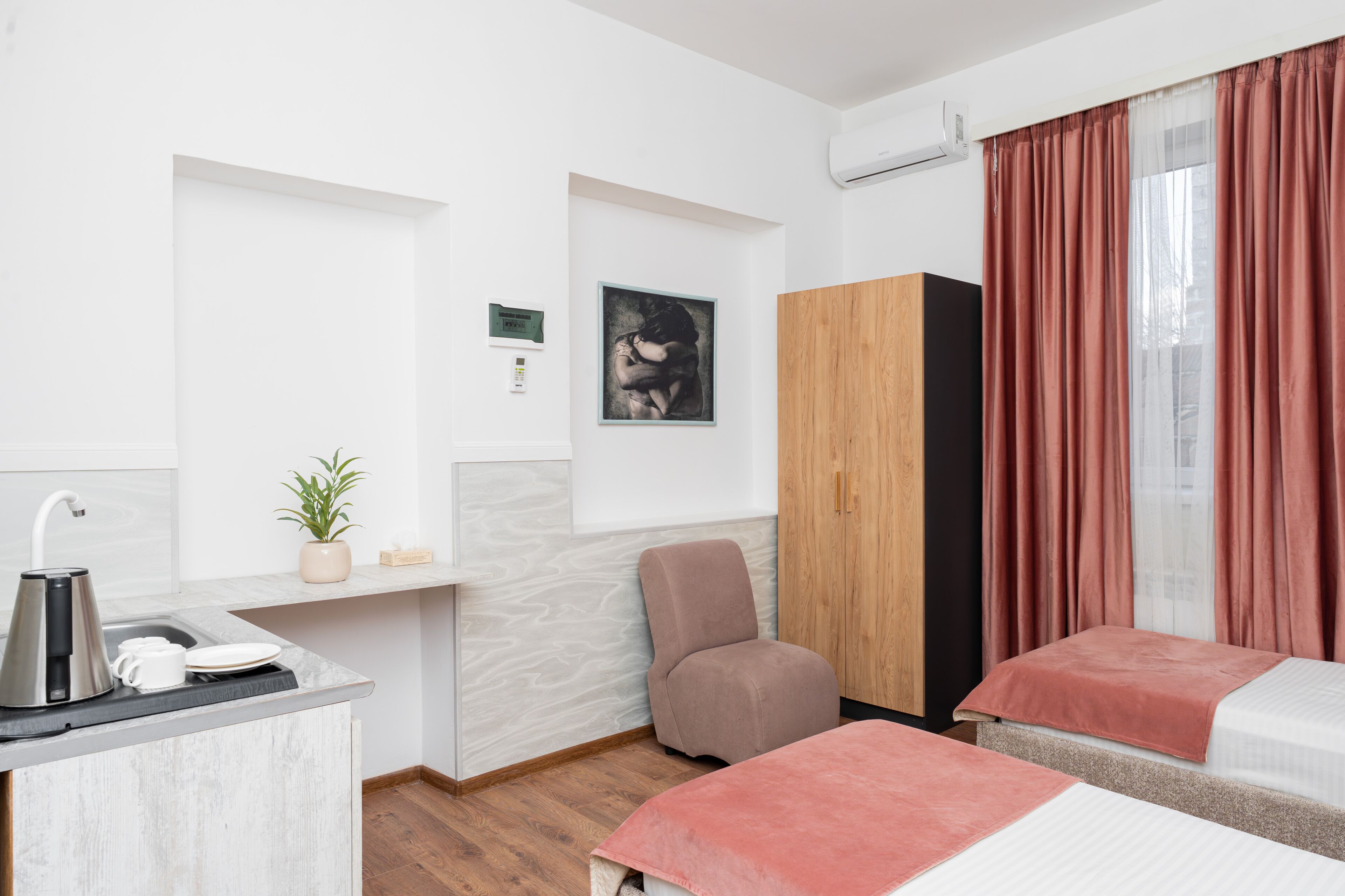 A hotel room with a kitchenette, a single bed, a chair, a wardrobe, and a large window with curtains.